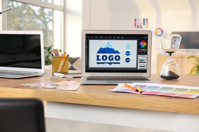 209505100_l A designer's workspace featuring a laptop displaying a mountain-themed logo design, color swatches, and an hourglass on a wooden desk.